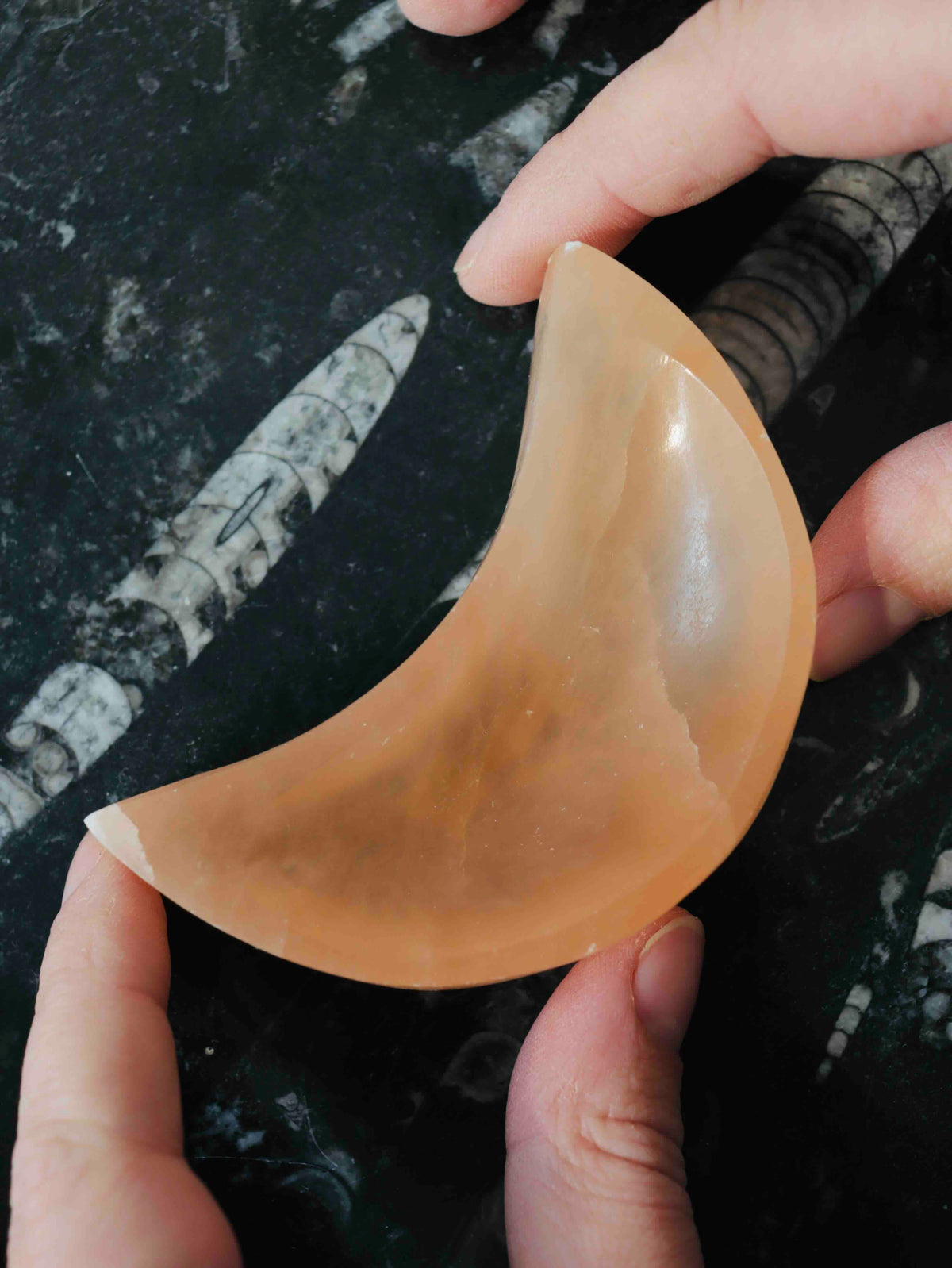 Mini bol en sélénite orange en forme de croissant de lune, tenu entre deux mains, montrant son intérieur creusé et ses reflets satinés.