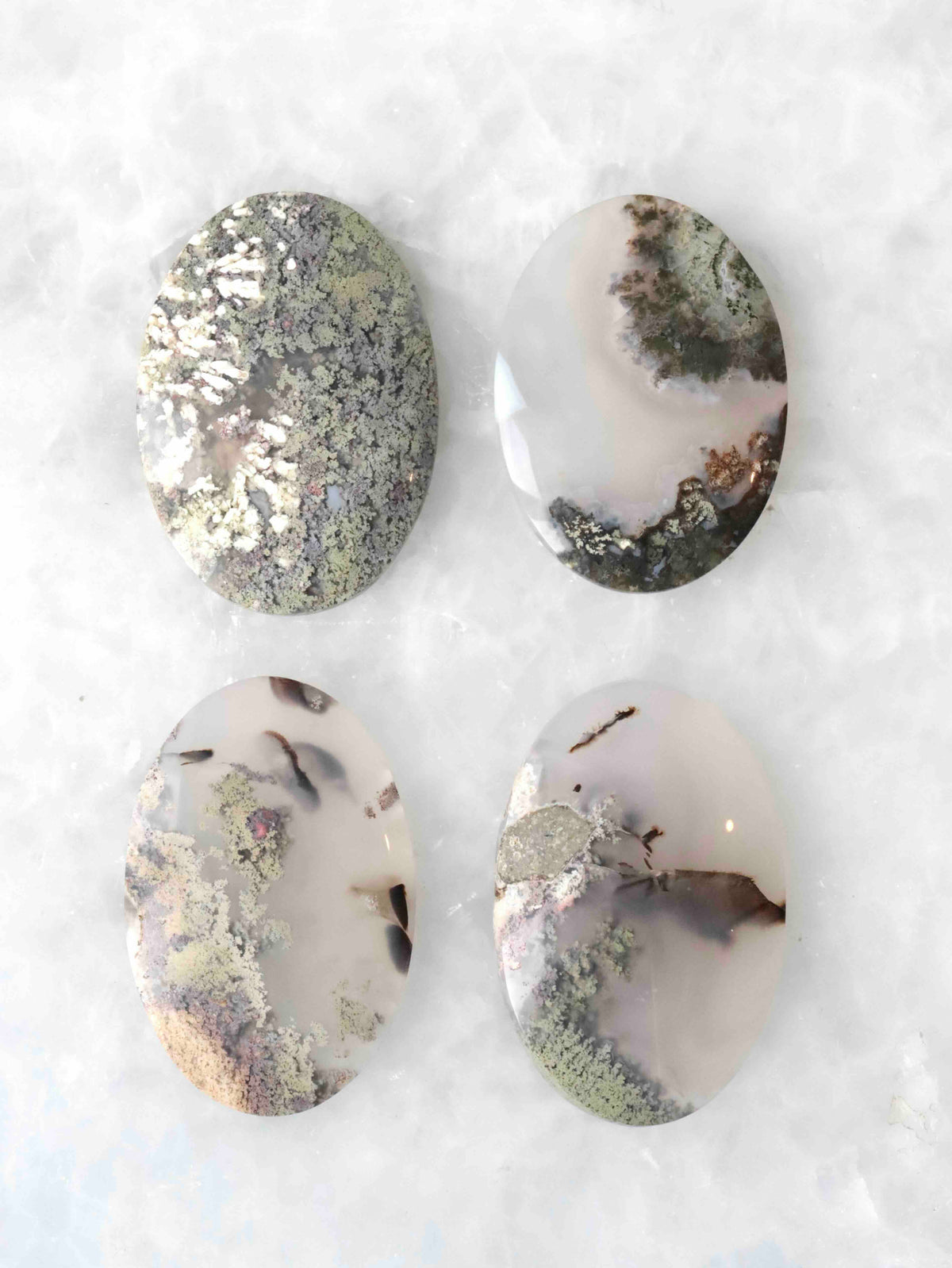 Quatre cabochons ovales polis en agate mousse, aux nuances blanc laiteux et translucides avec des inclusions vertes et brunes évoquant des végétaux, sur fond blanc.
