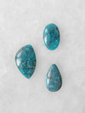 Trois cabochons polis en apatite bleue aux nuances turquoise et inclusions sombres, de formes ovale, libre et en goutte, sur fond clair.