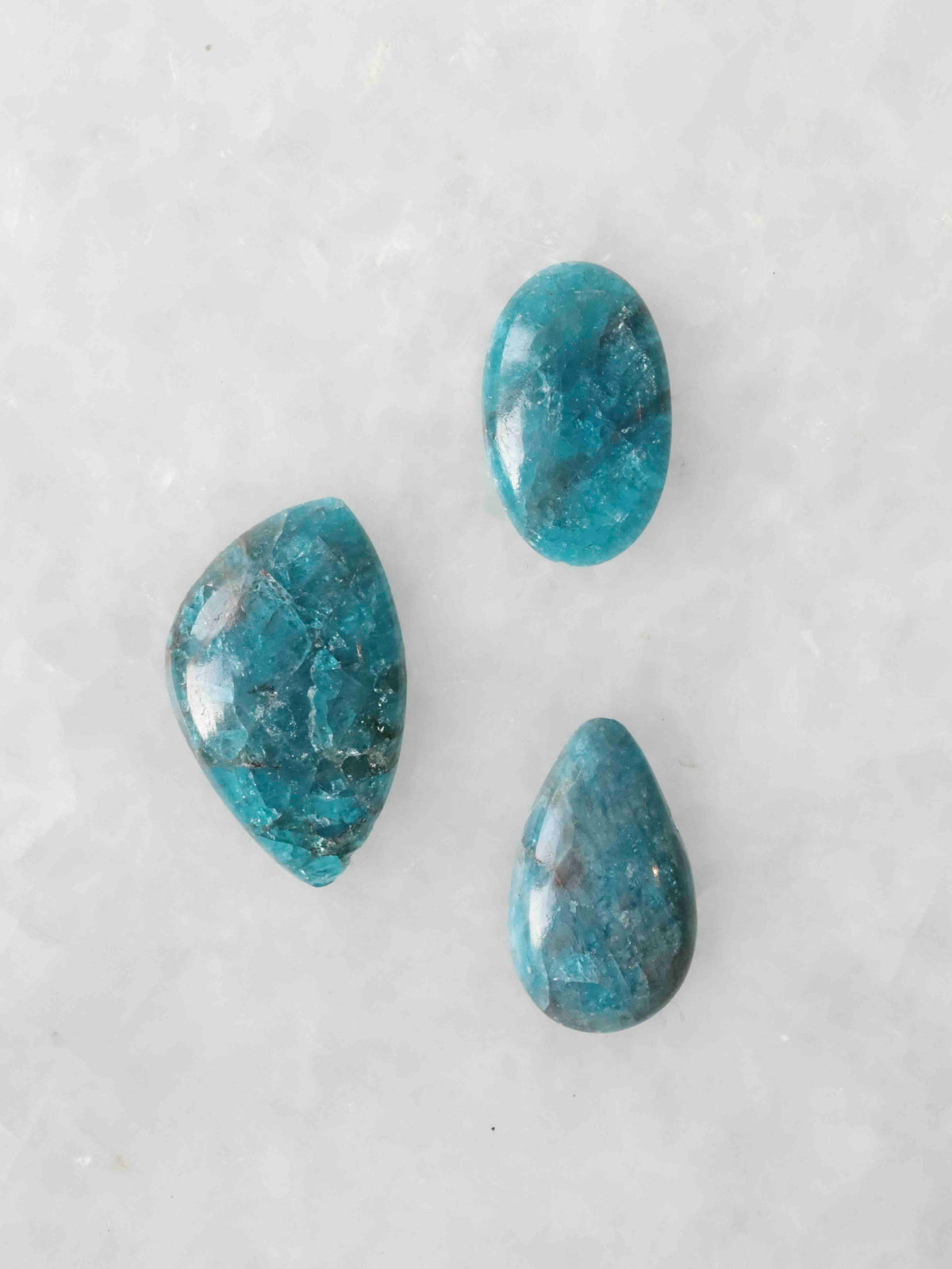 Trois cabochons polis en apatite bleue aux nuances turquoise et inclusions sombres, de formes ovale, libre et en goutte, sur fond clair.
