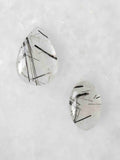 Deux cabochons polis de quartz transparent à inclusions de tourmaline noire en aiguilles, l’un en goutte et l’autre ovale, sur fond clair.
