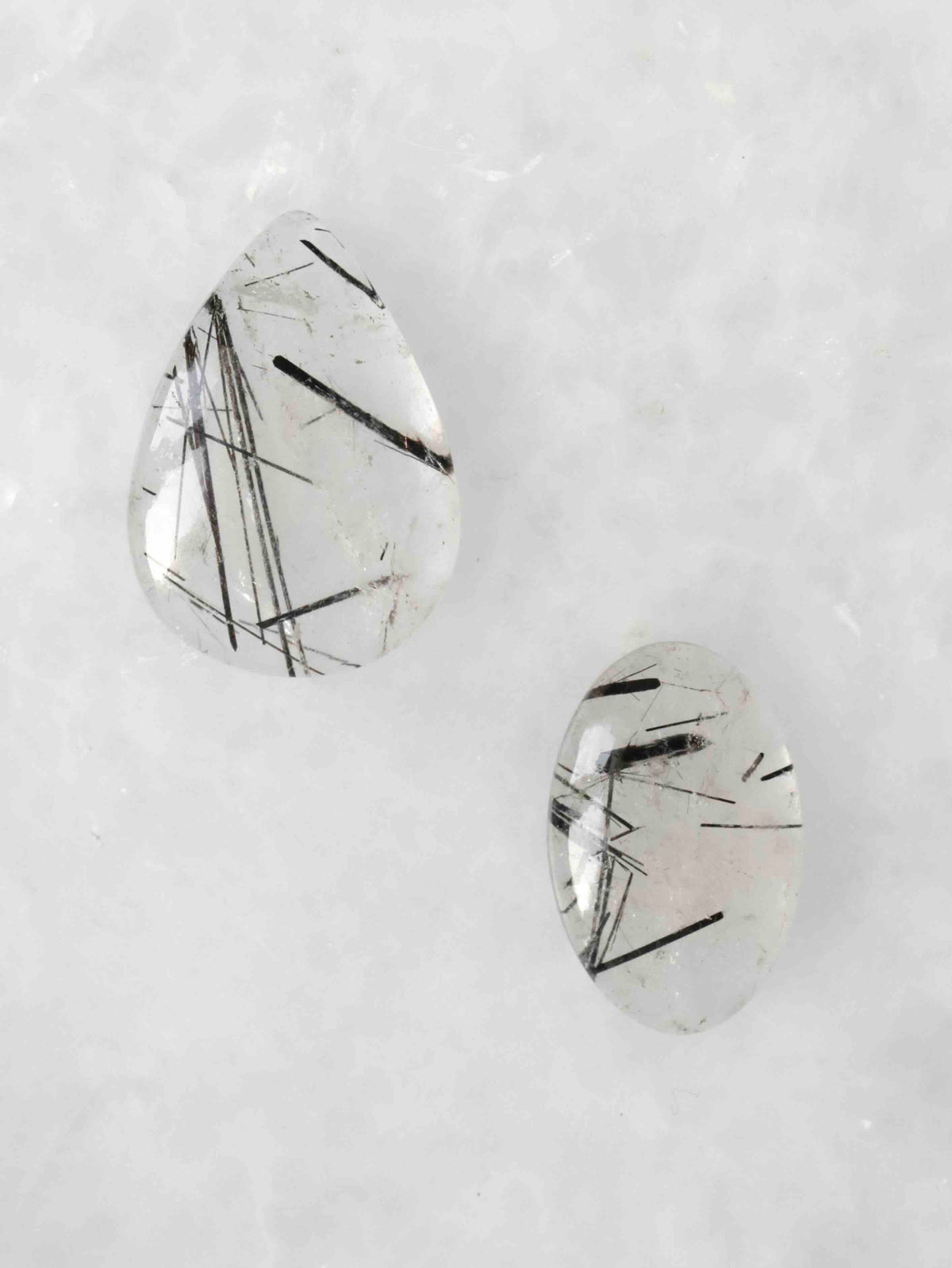 Deux cabochons polis de quartz transparent à inclusions de tourmaline noire en aiguilles, l’un en goutte et l’autre ovale, sur fond clair.