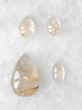 Quatre cabochons polis de quartz rutile translucide, en formes de goutte et ovale, traversés de fines aiguilles dorées, sur fond blanc.