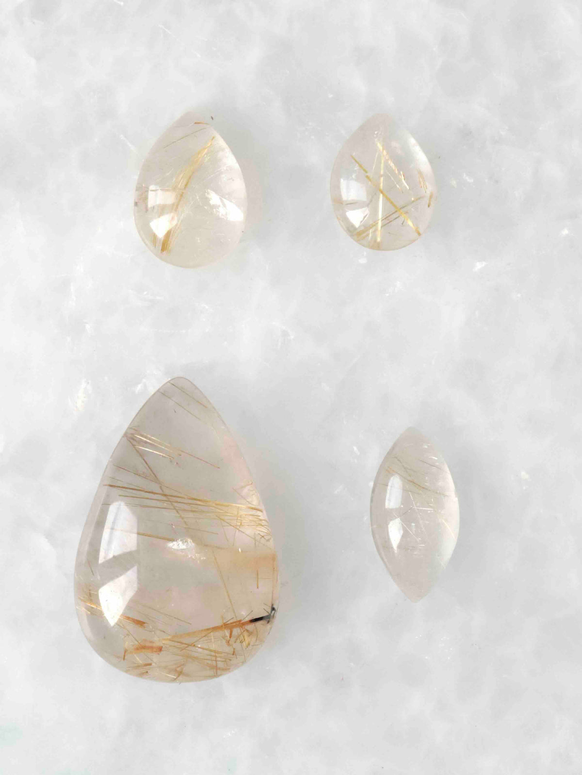Quatre cabochons polis de quartz rutile translucide, en formes de goutte et ovale, traversés de fines aiguilles dorées, sur fond blanc.