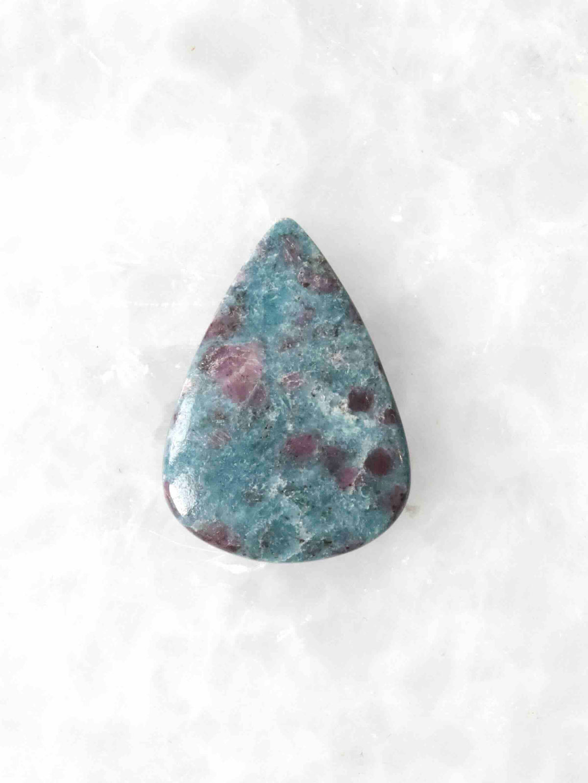 Cabochon poli en forme de goutte associant kyanite, fuchsite et rubis, aux nuances bleu-vert parsemées d’inclusions roses et pourpres, sur fond blanc.