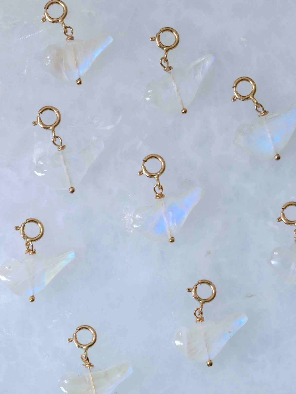  Charms oiseaux en pierre de lune aux reflets irisés, avec fermoirs en gold-filled 14K, disposés sur fond clair.