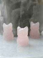 Chat sculpté en quartz rose vu de face, avec deux autres chats sur une surface claire en arrière-plan.