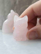 Petit chat sculpté en quartz rose pâle, vu de face et maintenu entre les doigts.