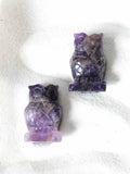 Deux chouettes sculptées en améthyste claire, pierre naturelle violette, vues de face sur fond blanc texturé