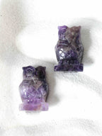 Deux chouettes sculptées en améthyste claire, pierre naturelle violette, vues de face sur fond blanc texturé