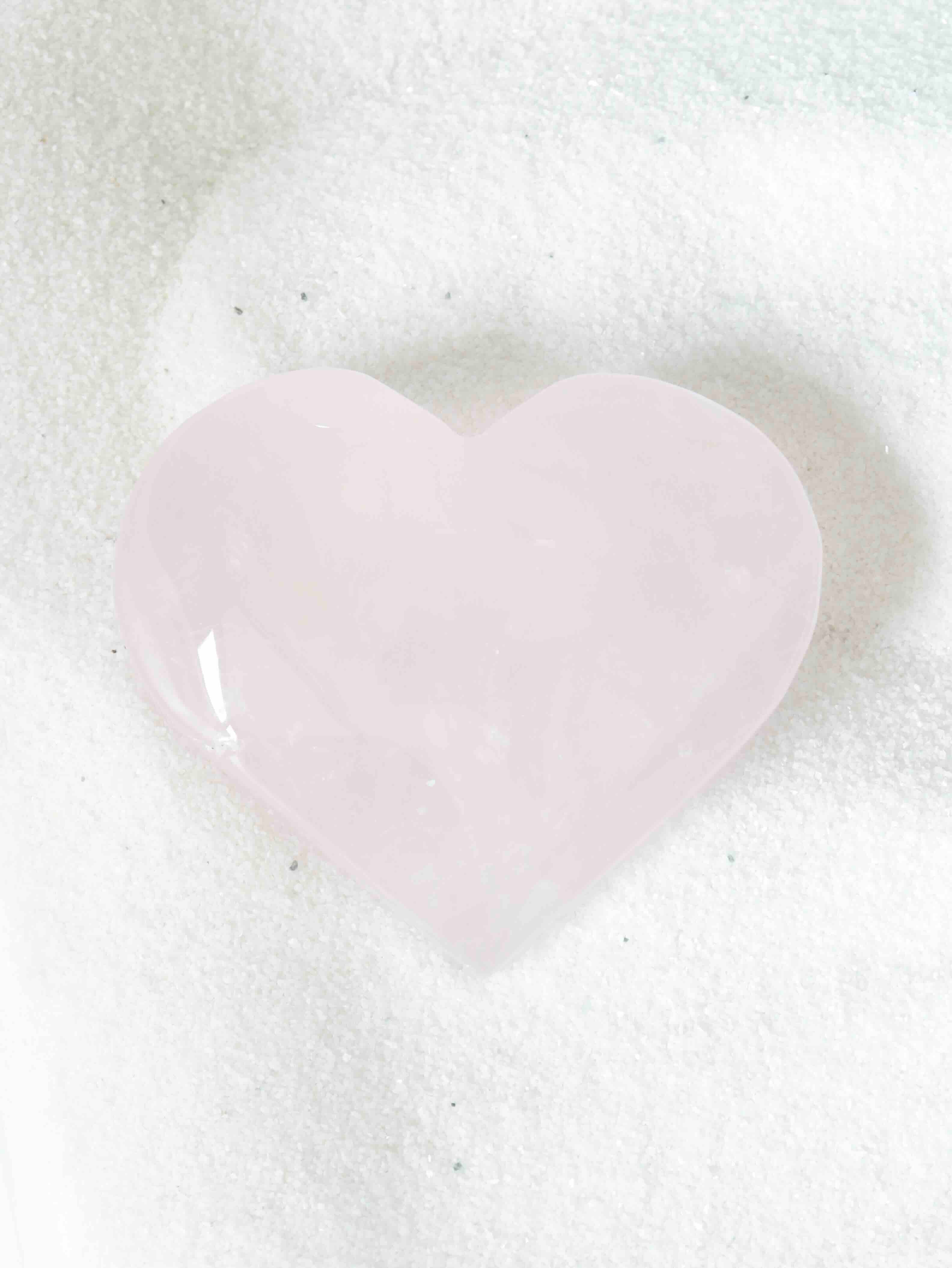 Cœur sculpté en quartz rose naturel, forme lisse et arrondie, posé sur fond blanc texturé
