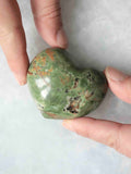  Cœur en chrysoprase verte polie, aux inclusions brunes et noires, tenu entre les doigts.
