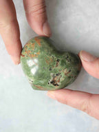  Cœur en chrysoprase verte polie, aux inclusions brunes et noires, tenu entre les doigts.