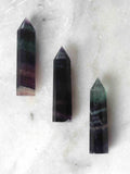 Petite pointe de fluorite multicolore semi-polie, nuances vert menthe et violet, posée sur marbre blanc