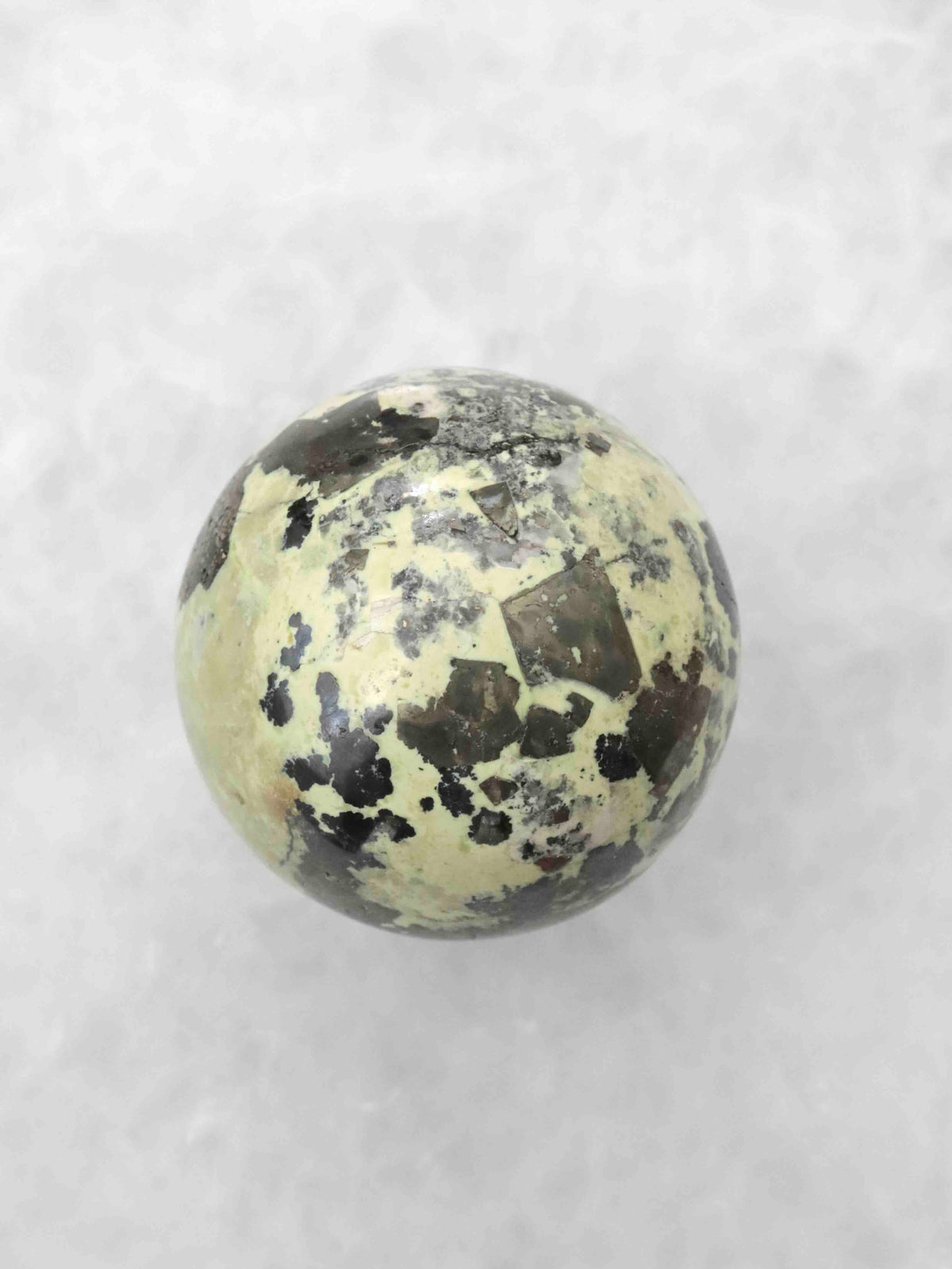 Sphère polie en serpentine vert pâle, avec de nombreux motifs anguleux noirs et gris répartis sur sa surface.