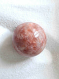 Sphère en pierre de soleil naturelle polie, teintes orange rosé et blanc nacré, vue de dessus sur fond blanc texturé