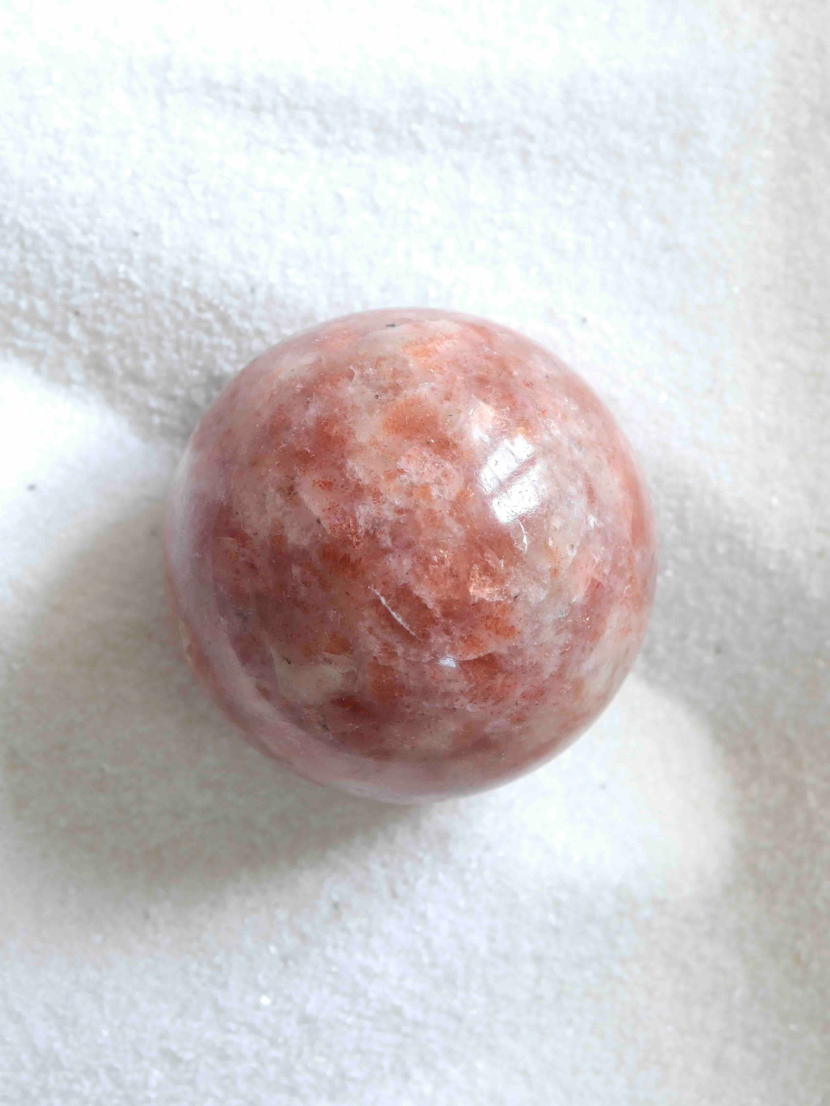 Sphère en pierre de soleil naturelle polie, teintes orange rosé et blanc nacré, vue de dessus sur fond blanc texturé