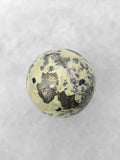 Sphère polie en serpentine vert pâle, parsemée de motifs noirs et gris aux reflets métalliques, sur fond blanc.