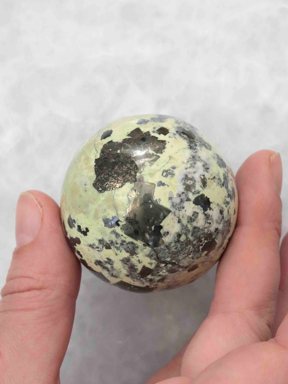Sphère en serpentine tenue entre les doigts, montrant ses nuances vert pâle et ses larges motifs sombres.